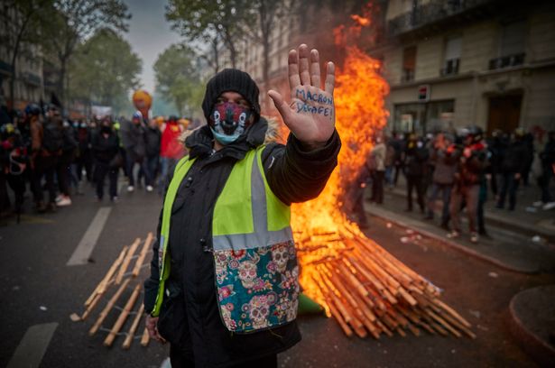 A Gilet Jaune, or yellow vest, protestor stands in front of a burning barricade holding his hand up with an inscription calling for President Macron to resign during a May Day protest in Paris