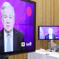 U.N. Secretary General Antonio Guterres (on screen), Prime Minister Yoshihide Suga (back) and Foreign Minister Toshimitsu Motegi attend an online summit co-hosted by Japan and Gavi, an alliance of governments, international organizations, companies and charities, on Wednesday night. | CABINET SECRETARIAT OF JAPAN / VIA KYODO