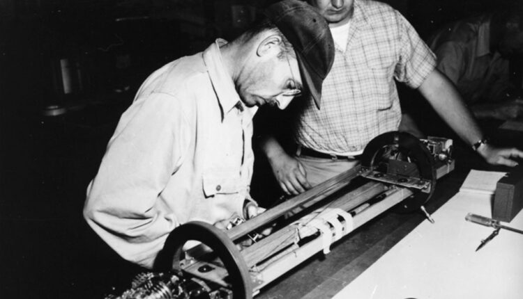 black and white photo of a man in a cap with glasses leaning over a tabletop device while another man looks on