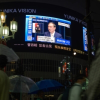 Pedestrians walk past a screen broadcasting a news conference by Prime Minister Yoshihide Suga in Tokyo on Thursday. | BLOOMBERG