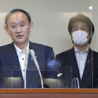 Prime Minister Yoshihide Suga speaks to reporters in Tokyo on Monday, as health minister Norihisa Tamura stands behind him. | POOL / VIA KYODO