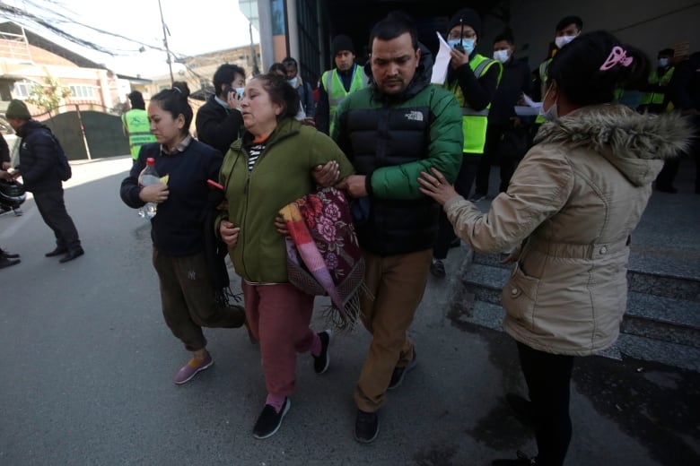 About a dozen people are visible with some buildings behind them. A woman is being held by the arm.