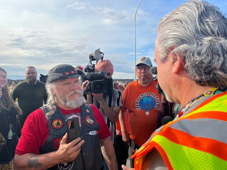A man holding a cellphone is shown speaking to a man in a neon vest. Four people stand behind him.