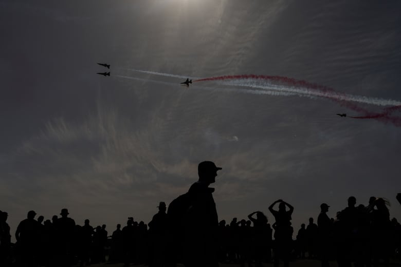 Spectators in silhouette watch aircraft performing in the sky, with colourful contrails.