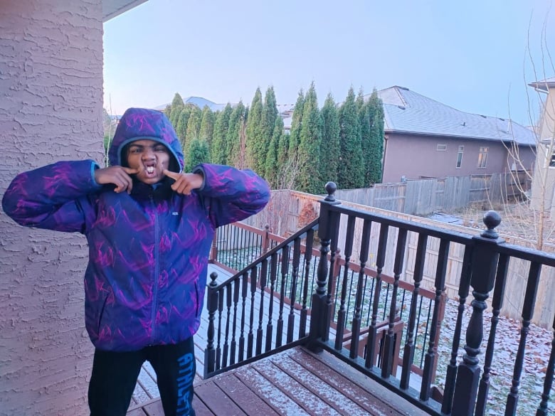 A young girl in a blue jacket sticks her tongue out and crosses her eyes at the back deck of a house.