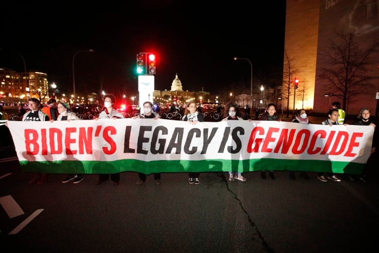 A line of people stand in on a street, with the U.S. Capitol building in the distance, holding a banner with the words "BIDEN'S LEGACY IS GENOCIDE" written in red and black letters on a white background with a green stripe below.