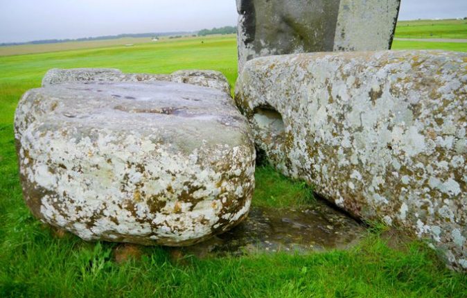 In this closeup photo, two mottled stones from Stonehenge lay on top of a darker brown stone that is embedded in the grass. That partially buried stone is the Altar Stone.