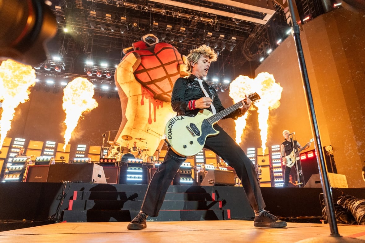 A male musician playing guitar on stage, with pyrotechnics erupting in the back of the stage.