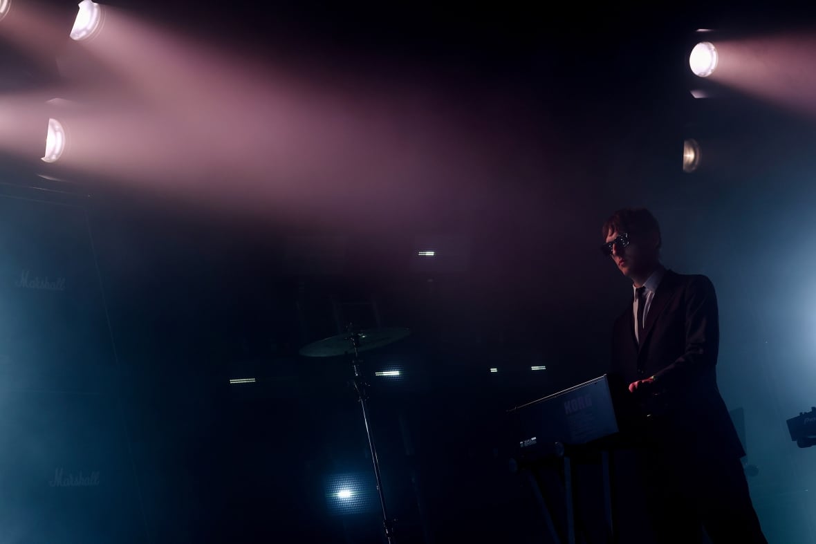 Several stage lights shining on stage as a male musician wearing a suit and dark sunglasses performs on stage.