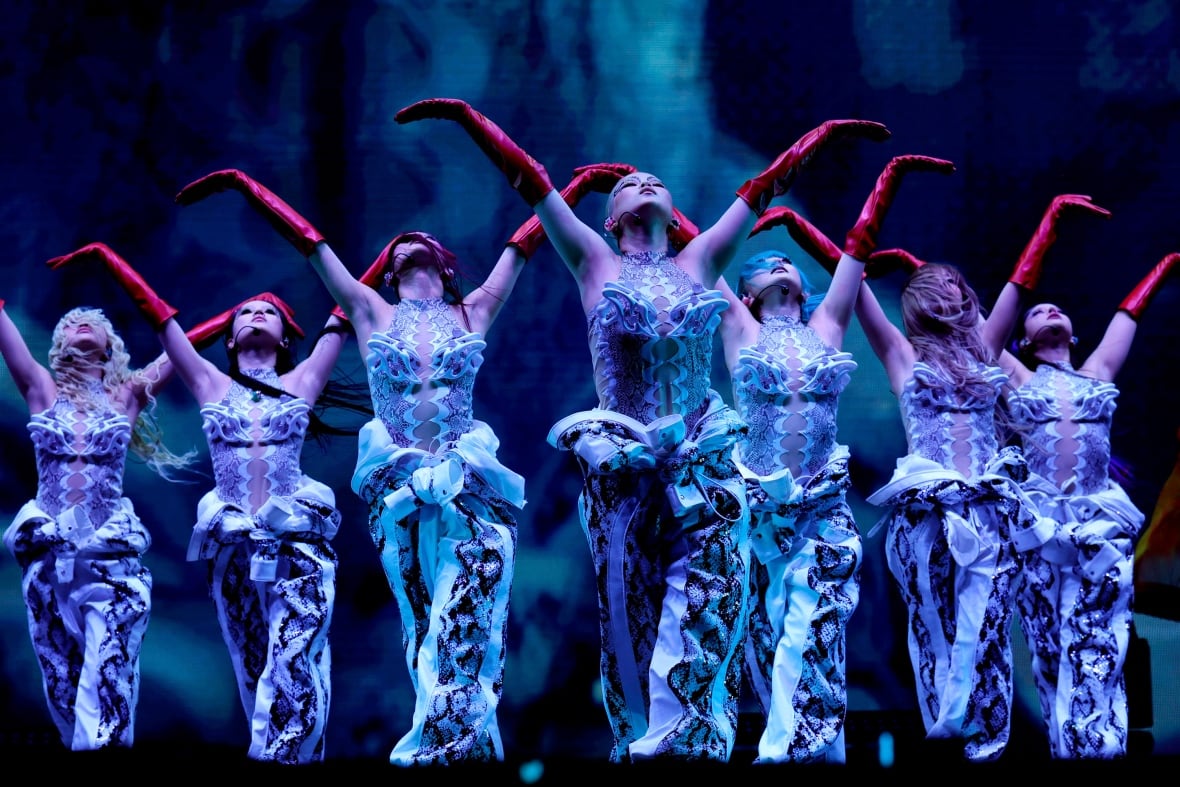 Female performers in costumes performing wearing red gloves with their hands up in the air in the shape of the letter V.