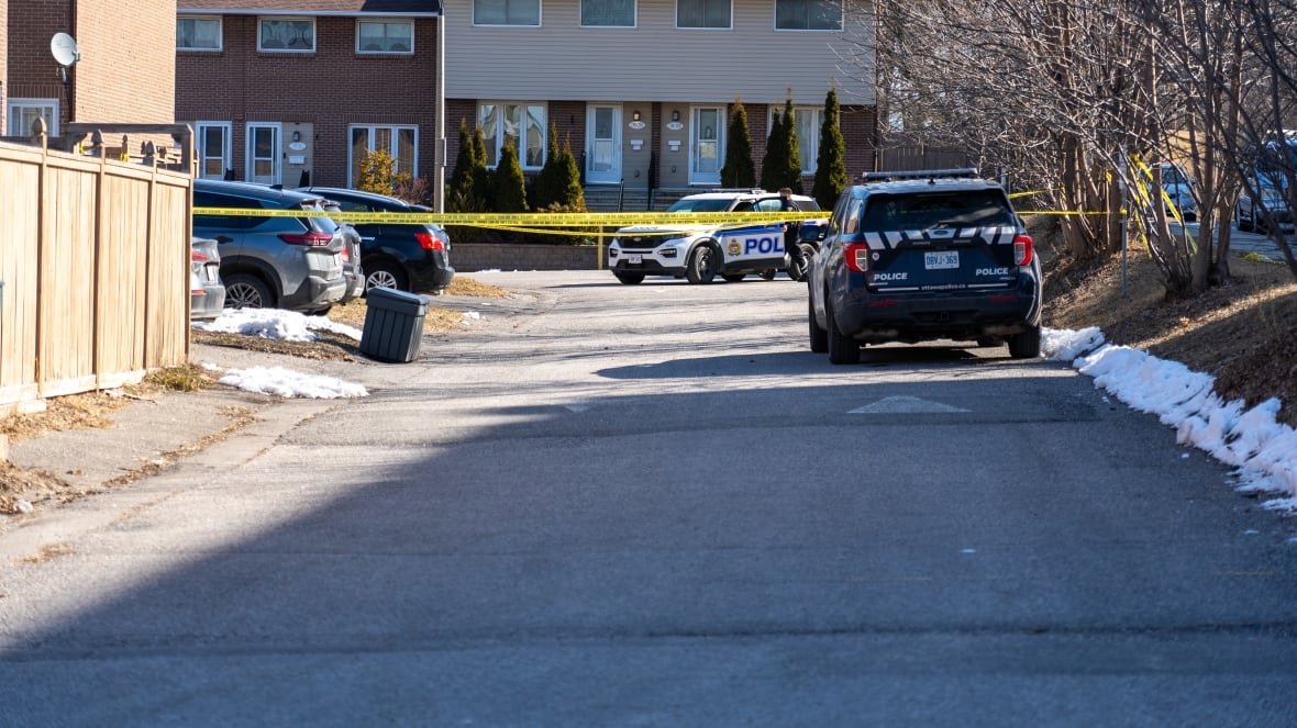 A photo of beige townhouses on a sunny day with a police car in the foreground. Some snow lingers on the pale grass. A few of the houses are marked off by police tape.