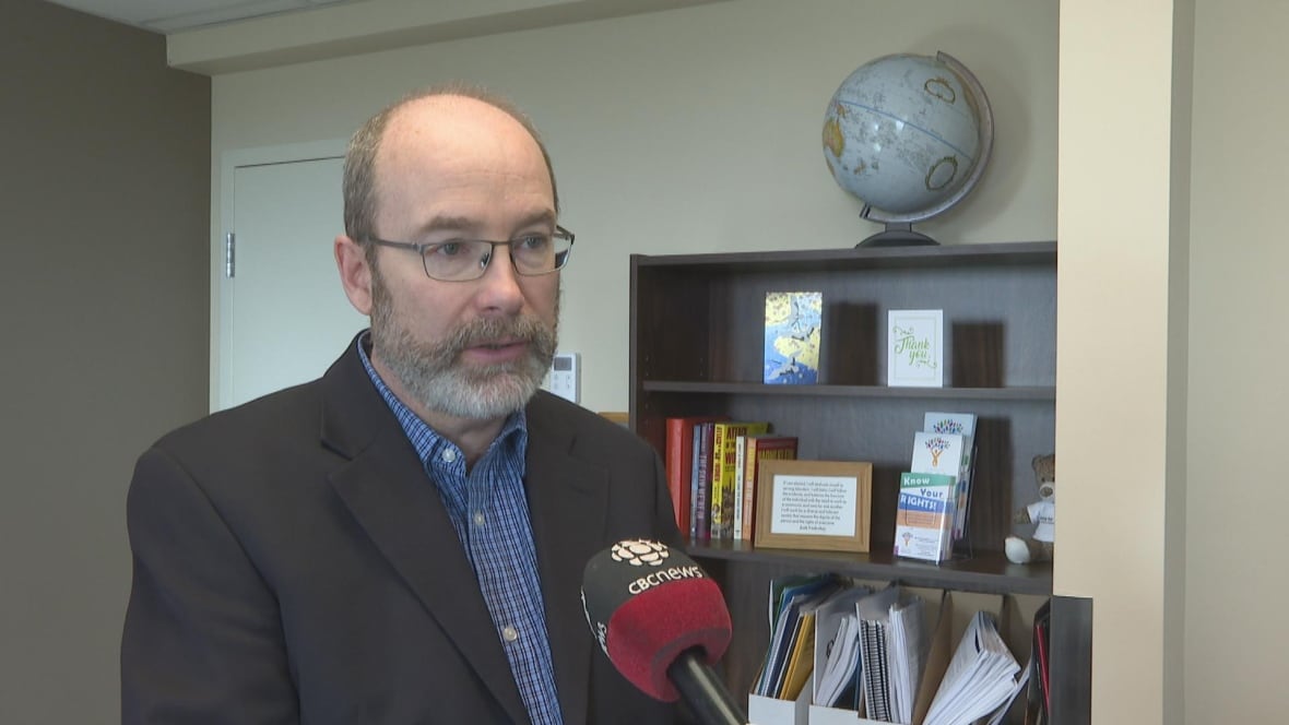 Man with glasses stand for an interview inside of an office. He is wearing a blazer and dress shirt. 