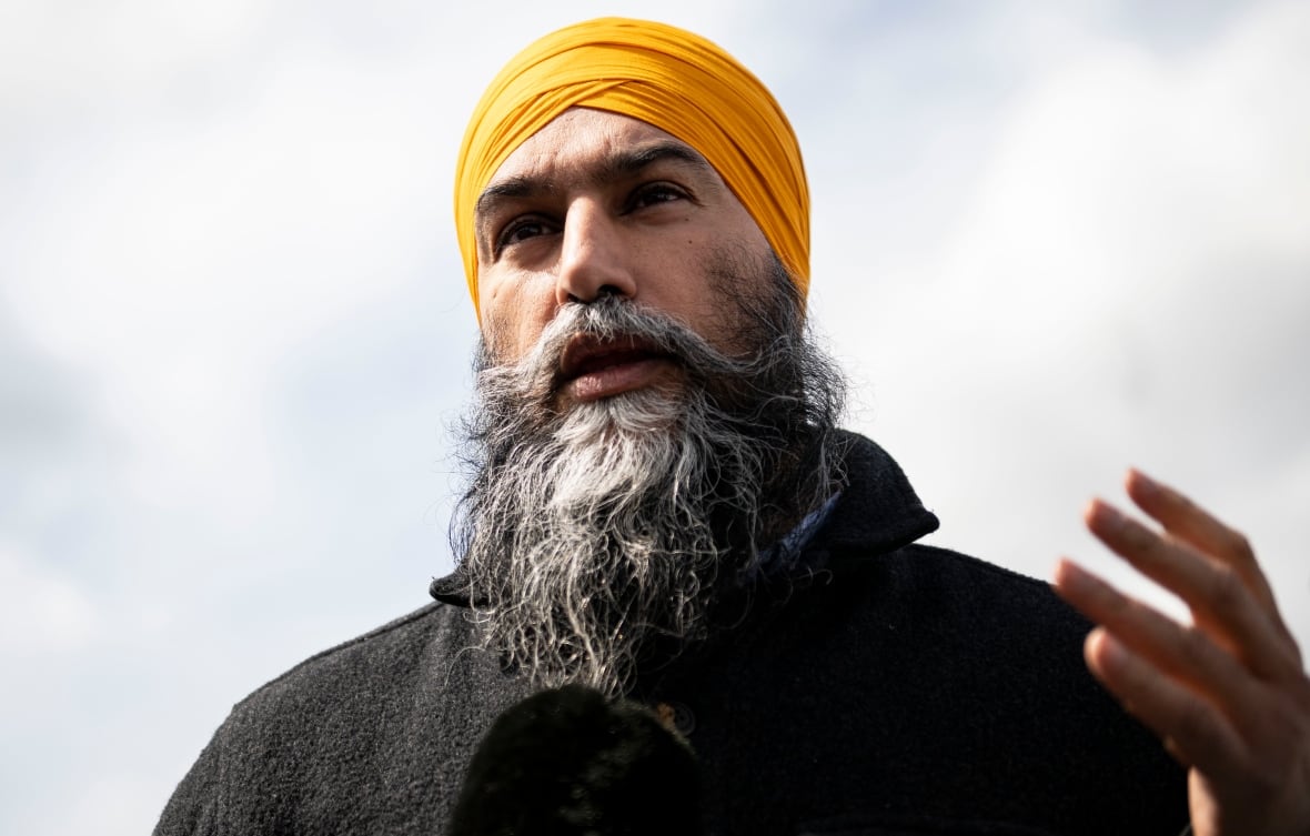 A man with a long beard and a yellow turban gestures while looking at the camera.