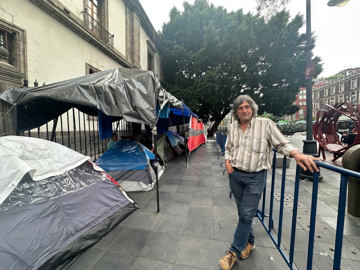 A man stands between metal fencing and a row of small tents.