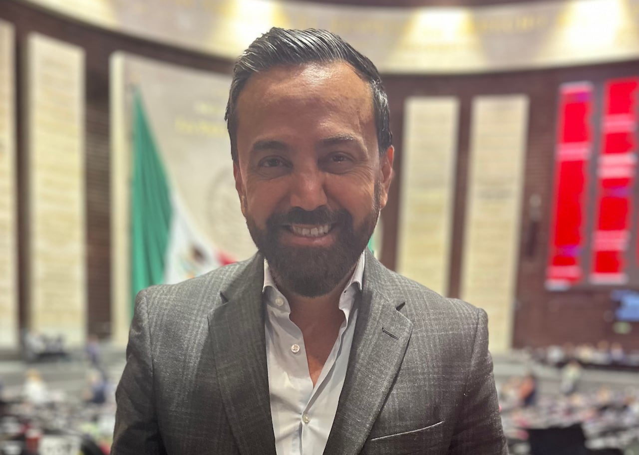 A man in a suit looks into the camera while standing in a wide chamber,