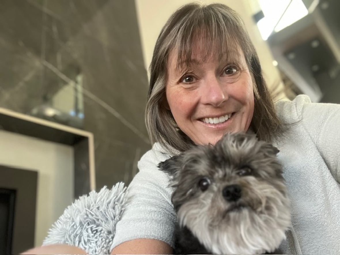 A smiling woman with blond highlights in her hair poses for a selfie with a long-haired grey and white dog under her chin.