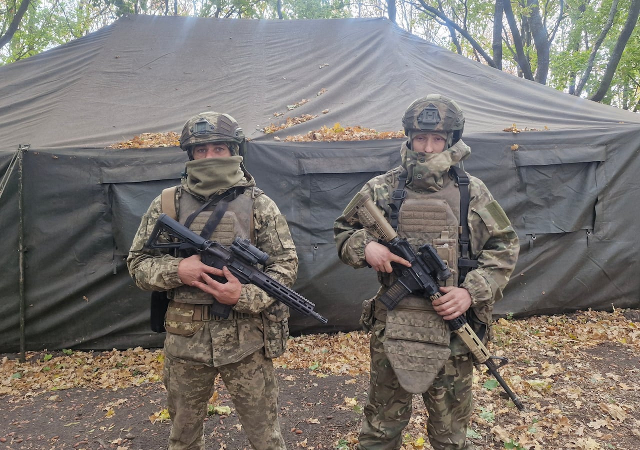 Two men in brown and green military fatigues, wearing vests and helmets, stand in front of a large black tent in a treed area. They each hold a rifle.