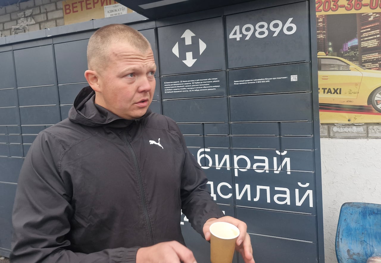 A man wearing a black jacket and holding a paper coffee cup gestures while talking next to a block of black mailboxes.