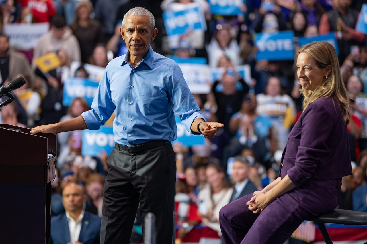 A dark-complected, cleanshaven man in a blue-collared shirt stands at a podium and points toward a blonde-haired woman in jacket and pants who is seated.