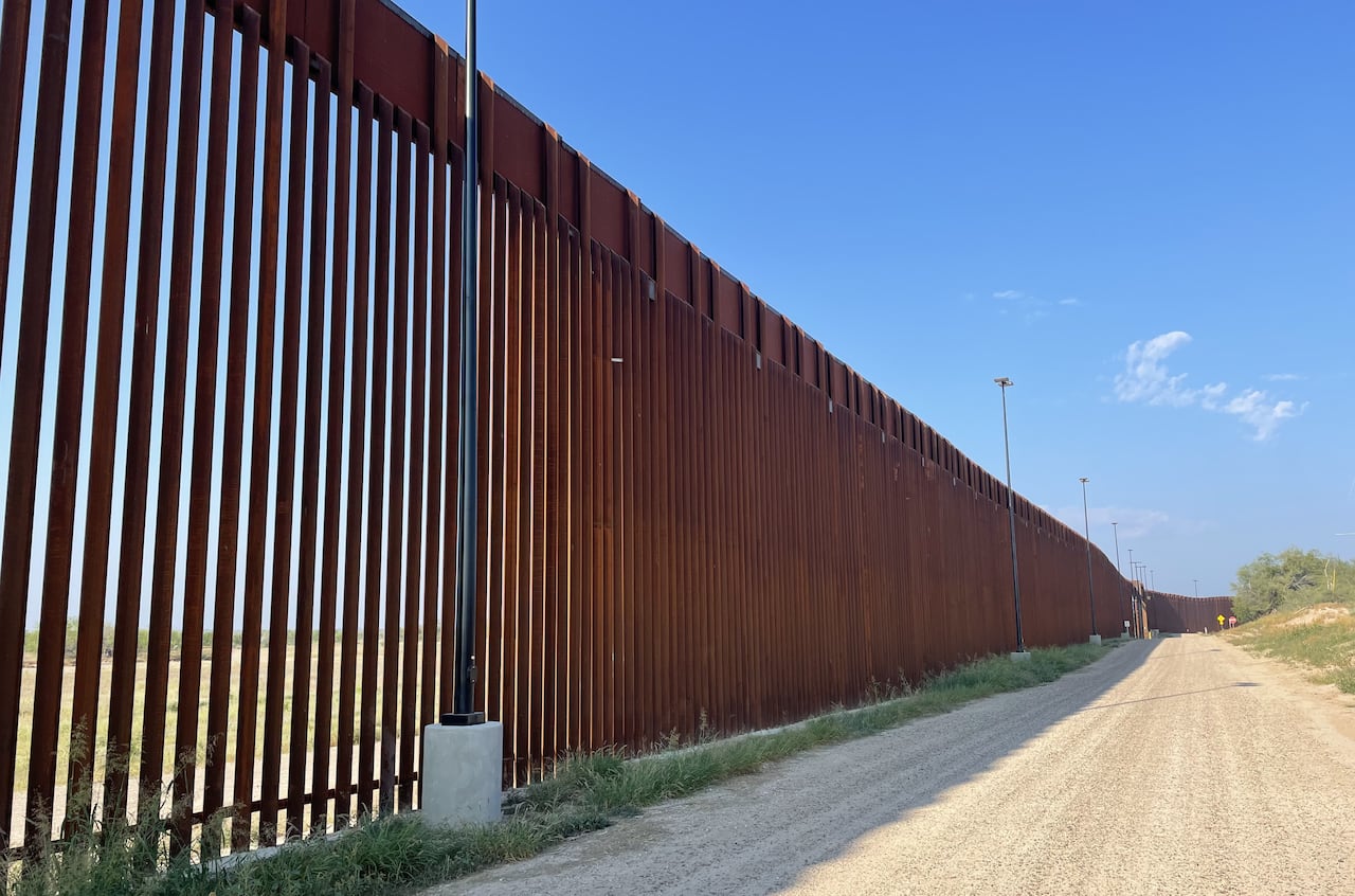 A tall metal fence stretches into the distance, along a dusty road.