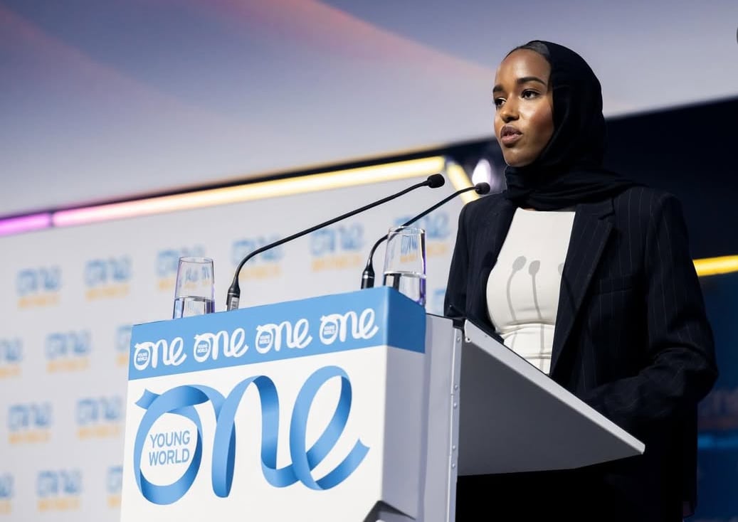 A woman wearing a black hijab speaks at a podium with a logo that says One Young World.