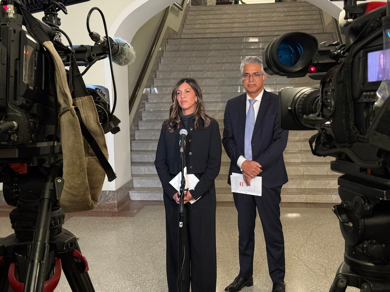 A man and a woman stand in front of a staircase at a microphone, with cameras facing them on either side.