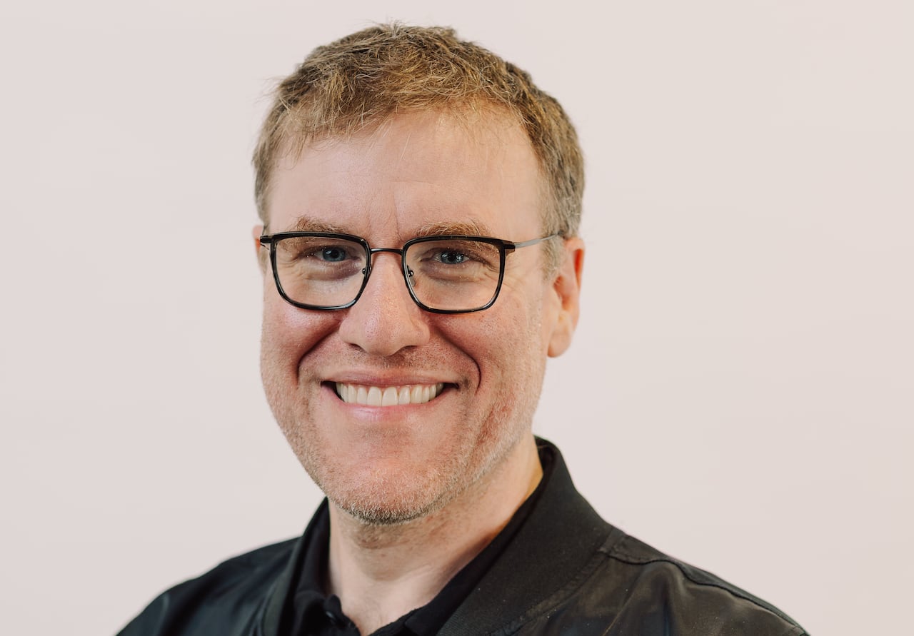 A smiling man with glasses wearing a black bomber jacket