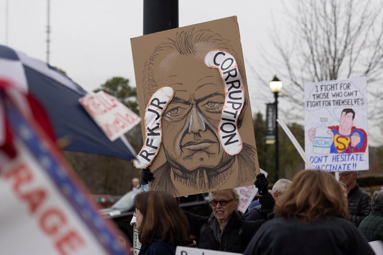 a drawing of politician RFK jr on a poster with worms coming out of his head. people in a crowd outside hold signs with pro vaccine messages in protest