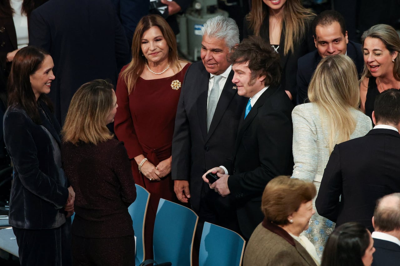 Several people stand near seats, men and women, in formal wear at an apparent ceremony.