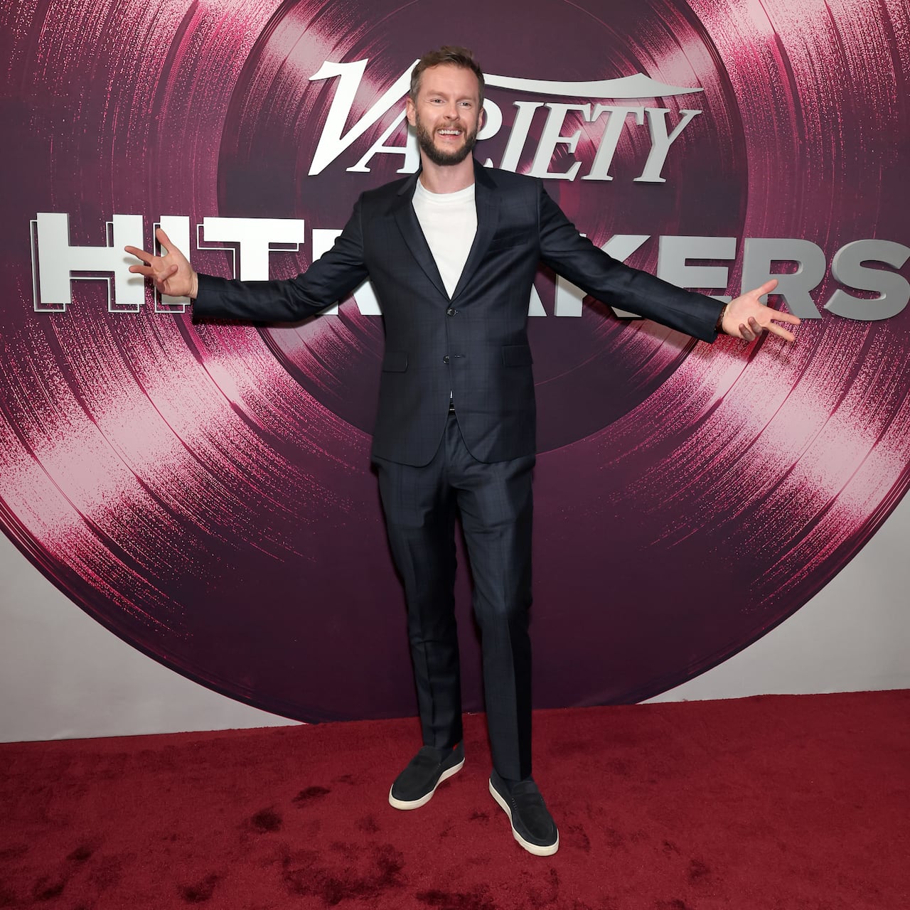 A thin, bearded man holds his hands out as his stands in front of a giant record.