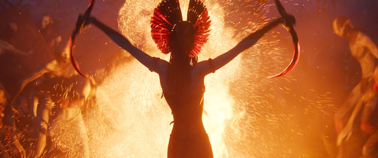 A computer-generated image of an alien silhouetted in front of a large bonfire is shown. The alien is holding two sickle blades.