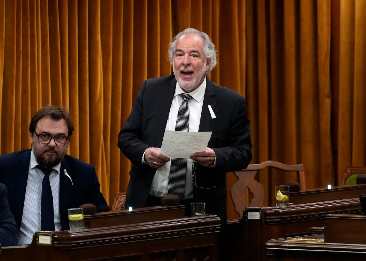 A man in a suit holds a piece of paper as he speaks in a legislature.