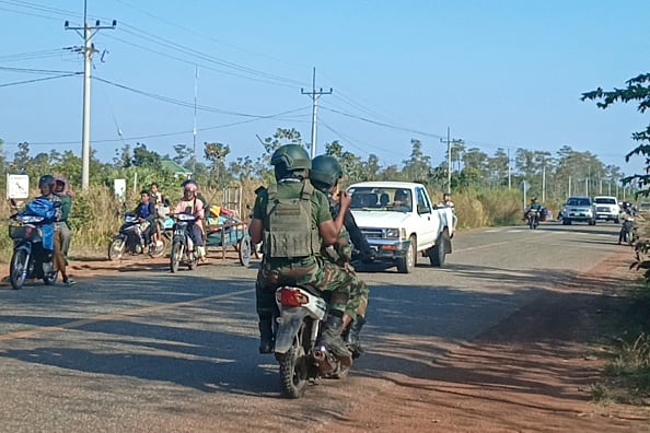 Two soldiers in uniform ride on an motorcyle, while people in cars and trucks and on bicycles ride in the opposite direction.