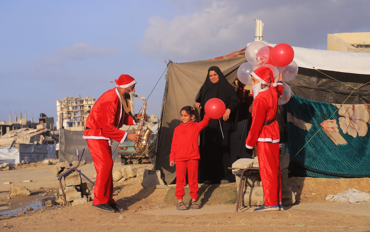 Two people dressed in Santa Claus costumes play saxophone and hold balloons as a child dressed in red smiles and holds a red balloon as a woman in black behind her looks on.