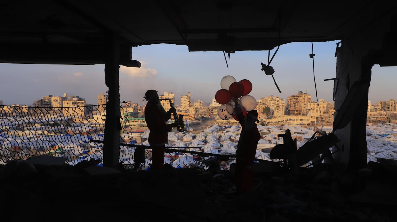 The silhouettes of a man playing saxophone and a boy holding balloons are seen through rubbles and a tent city is seen below.
