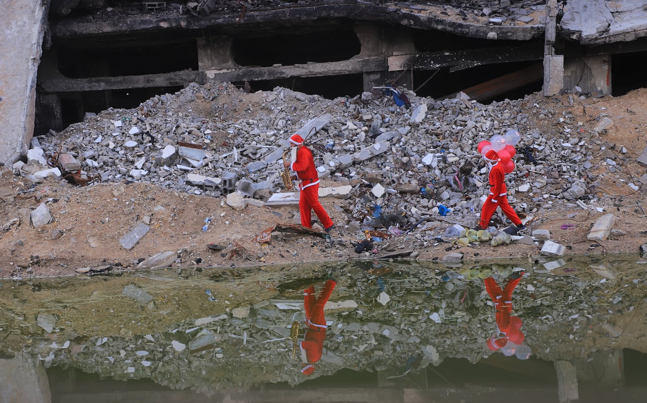 Two people dressed as Santa walk through rubble as a man plays a saxophone and a boy carries balloons.