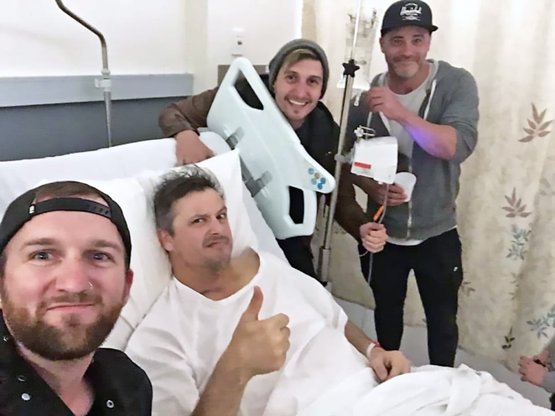Three men around male patient in hospital bed with thumbs up.